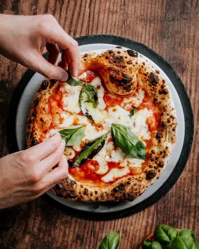 Home Close-up of hands adding basil to a freshly baked Neapolitan pizza, highlighting its cheesy and delicious appeal.